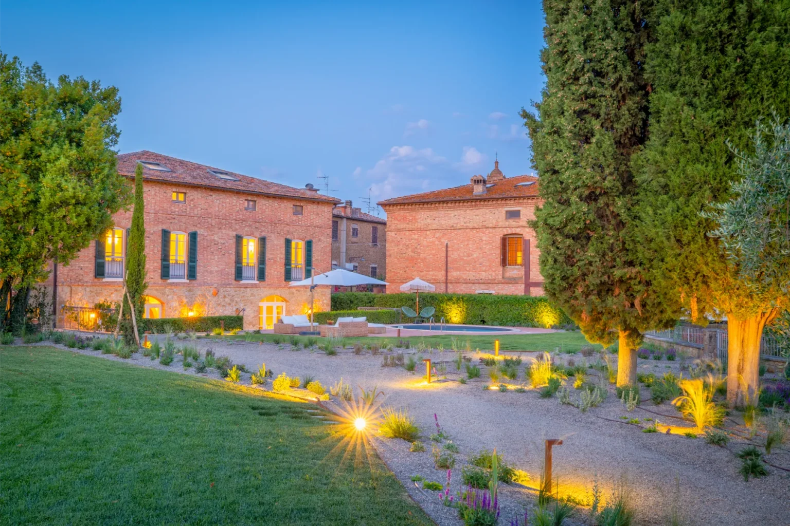 Beautifully landscaped garden featuring a rustic brick building, illuminated by soft lighting at dusk. Lush greenery, modern seating areas, and decorative plants enhance the serene outdoor space, perfect for relaxation. | Only on SopranoVillas