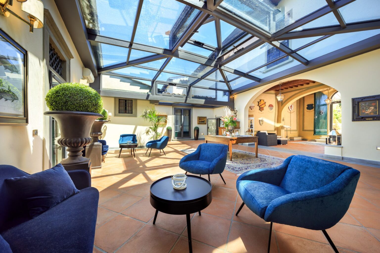 Bright and airy sunroom featuring a glass roof, stylish blue chairs, and decorative plants. The space includes a wooden table, artwork on the walls, and terracotta flooring, creating a modern yet cozy atmosphere. Ideal for relaxation and social gatherings. | Only on SopranoVillas