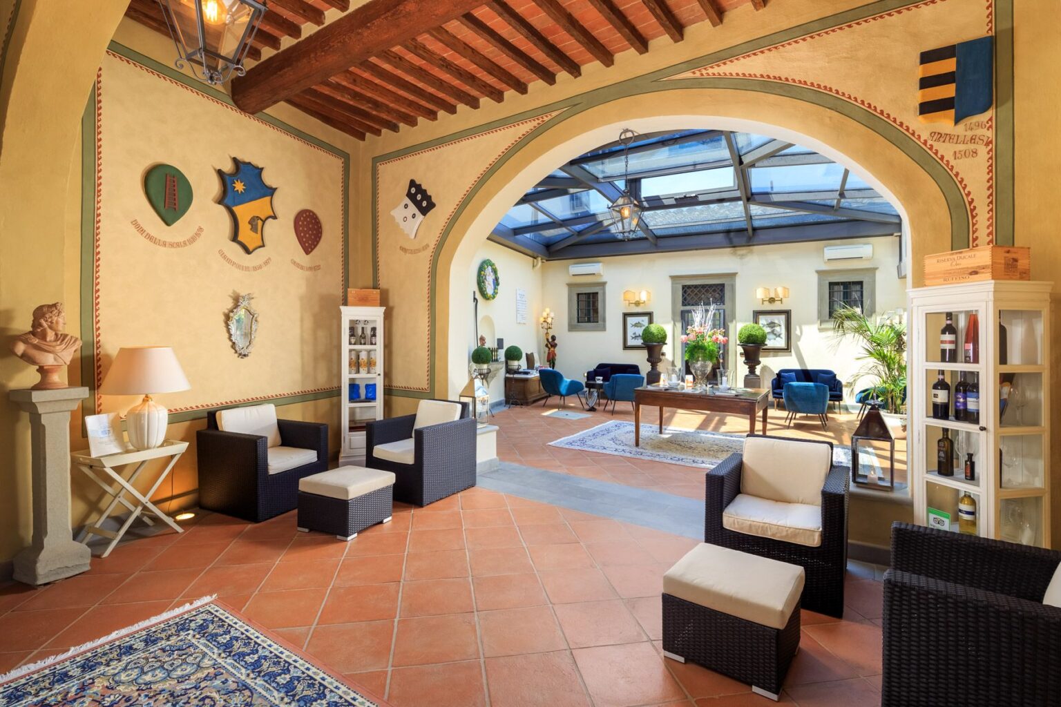 Cozy lounge area featuring modern wicker furniture, decorative shields on the walls, and a glass ceiling that allows natural light. The space includes a bookshelf displaying drinks, plants, and elegant decor, creating an inviting atmosphere for relaxation or social gatherings. | Only on SopranoVillas