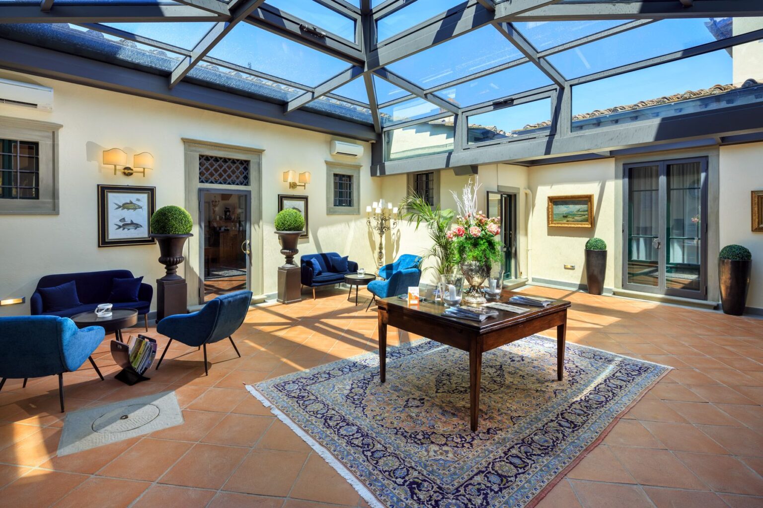 Bright and airy indoor lounge featuring a glass ceiling, elegant blue seating, a wooden table with decorative items, and potted greenery, creating a welcoming atmosphere. | Only on SopranoVillas