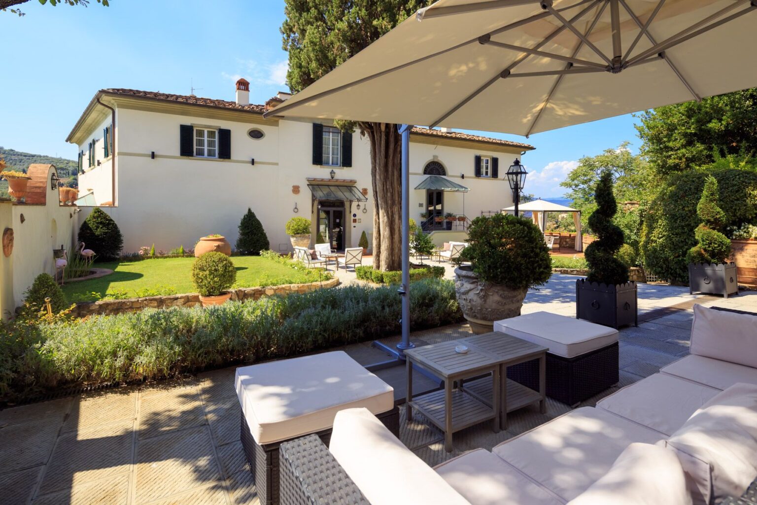 Elegant villa with a beautifully landscaped garden, featuring manicured hedges, potted plants, and outdoor seating areas. The image captures a sunny day with a clear blue sky, showcasing the villa's charming architecture and inviting outdoor spaces, perfect for relaxation and entertaining. | Only on SopranoVillas