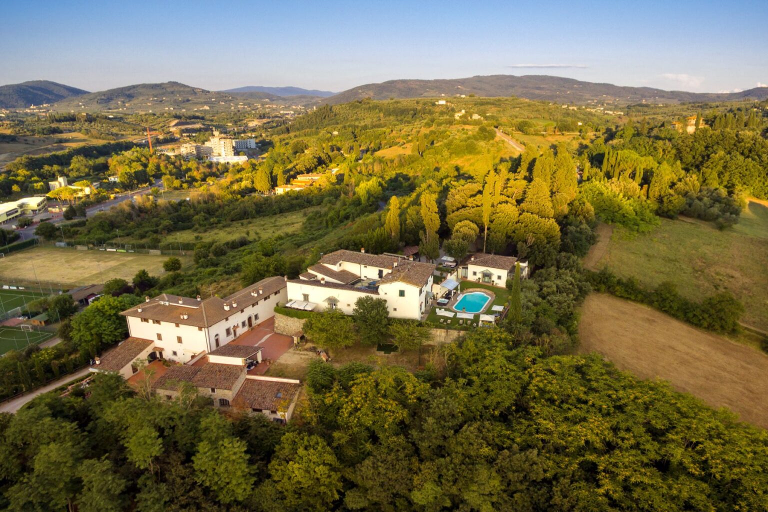 Aerial view of a serene countryside villa surrounded by lush greenery, featuring a swimming pool and rustic architecture, with rolling hills and distant mountains in the background. Ideal for vacation rentals and nature retreats. | Only on SopranoVillas