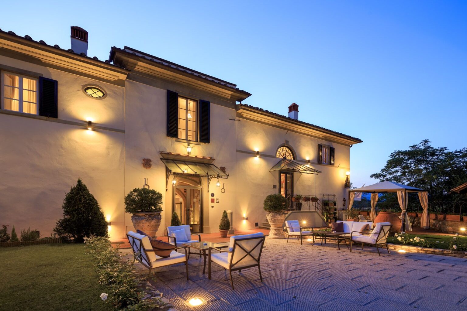 Elegant villa exterior at dusk, featuring a beautifully landscaped garden, outdoor seating area with stylish furniture, and warm lighting illuminating the facade. The image showcases architectural details, including windows with black shutters and a charming entrance. | Only on SopranoVillas