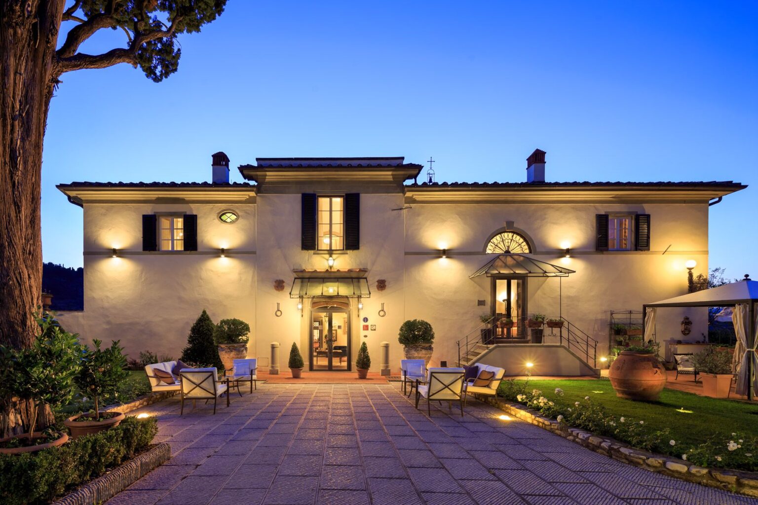 Elegant villa at dusk featuring warm exterior lighting, a landscaped entrance with seating, and a charming staircase leading to the main door, surrounded by greenery and decorative pottery. | Only on SopranoVillas