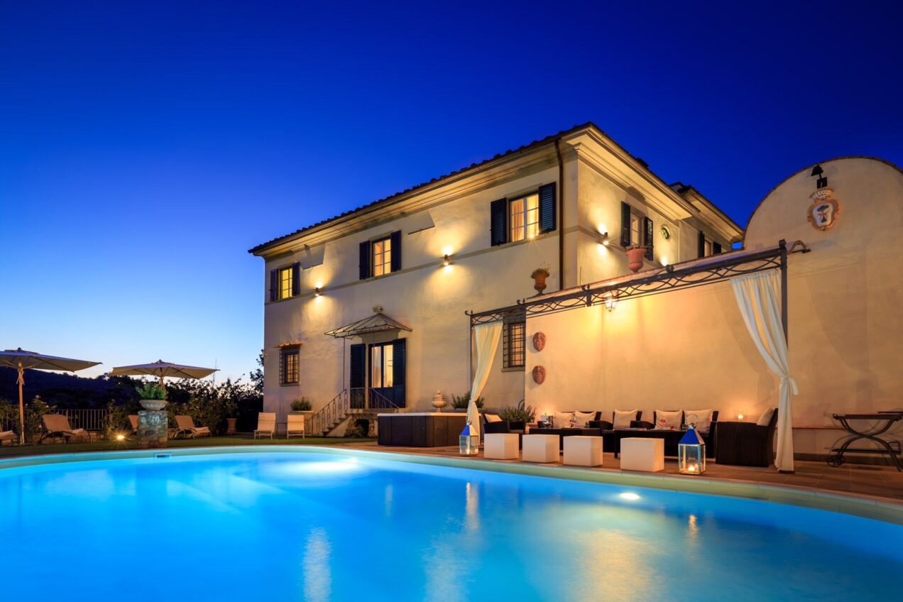 Luxurious villa with a sparkling blue swimming pool, elegant outdoor seating area, and beautifully lit facade, set against a twilight sky. Perfect for relaxation and outdoor gatherings. | Only on SopranoVillas