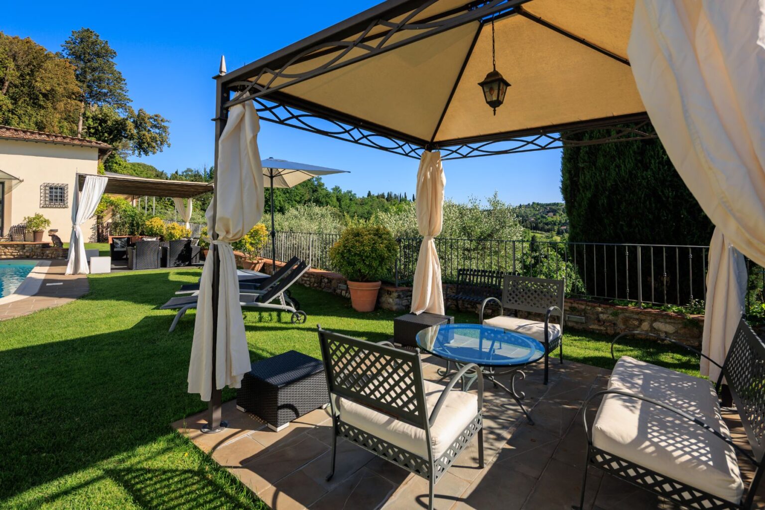Outdoor seating area featuring a gazebo with soft drapes, a round glass table, and comfortable chairs, surrounded by lush greenery and a swimming pool, ideal for relaxation and entertaining. | Only on SopranoVillas