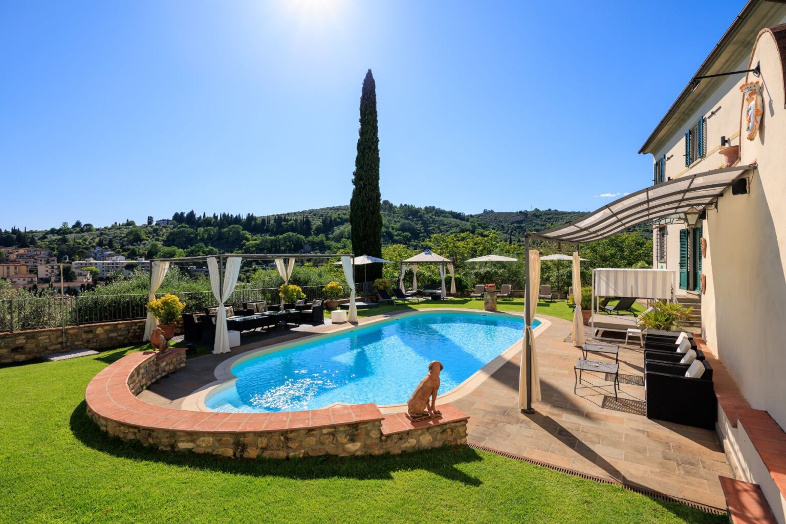 Luxury villa with a swimming pool surrounded by a beautifully landscaped garden, featuring lounge chairs and shaded areas, set against a backdrop of rolling hills and clear blue skies. Perfect for relaxation and outdoor gatherings. | Only on SopranoVillas