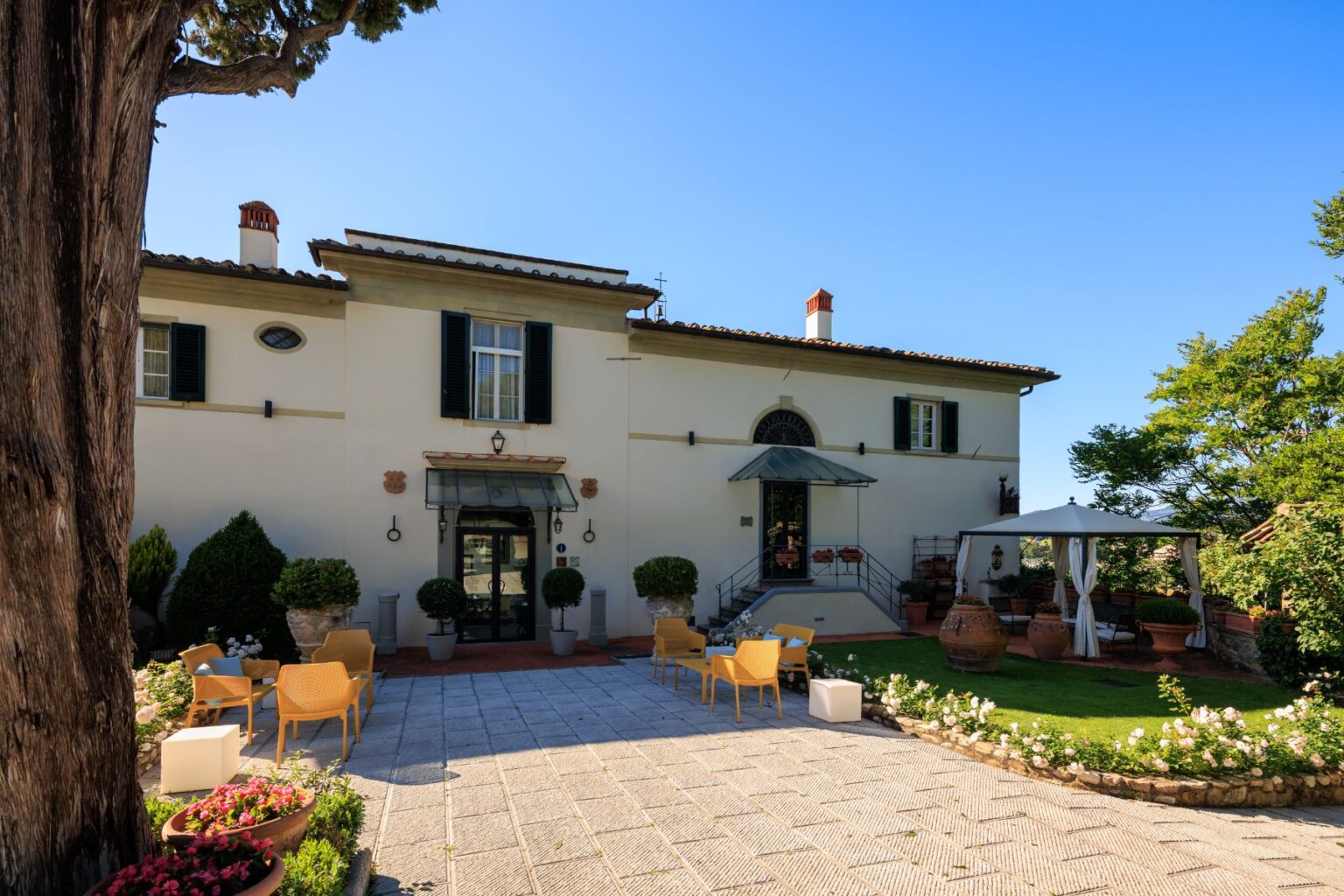 Elegant villa exterior featuring a charming courtyard with seating, surrounded by lush greenery and vibrant flowers, under a clear blue sky. Ideal for outdoor gatherings and relaxation. | Only on SopranoVillas