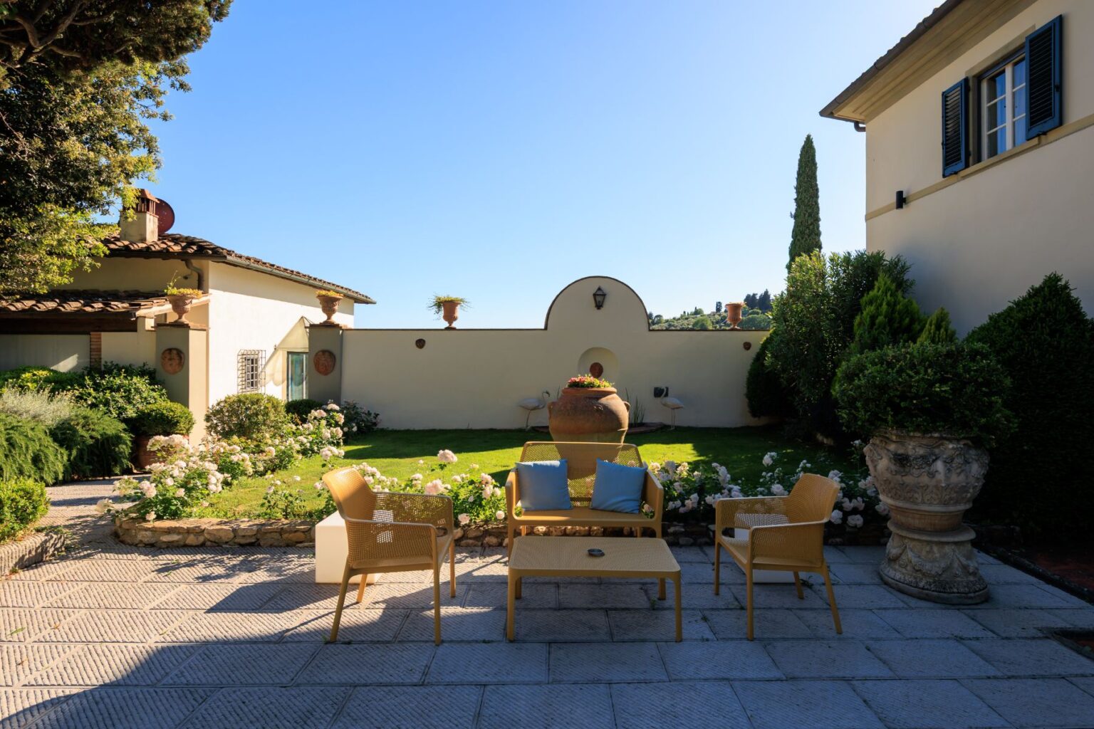Cozy outdoor seating area with yellow chairs and a coffee table, surrounded by lush greenery and blooming roses, set against a clear blue sky and a charming wall. Ideal for relaxation and enjoying nature. | Only on SopranoVillas