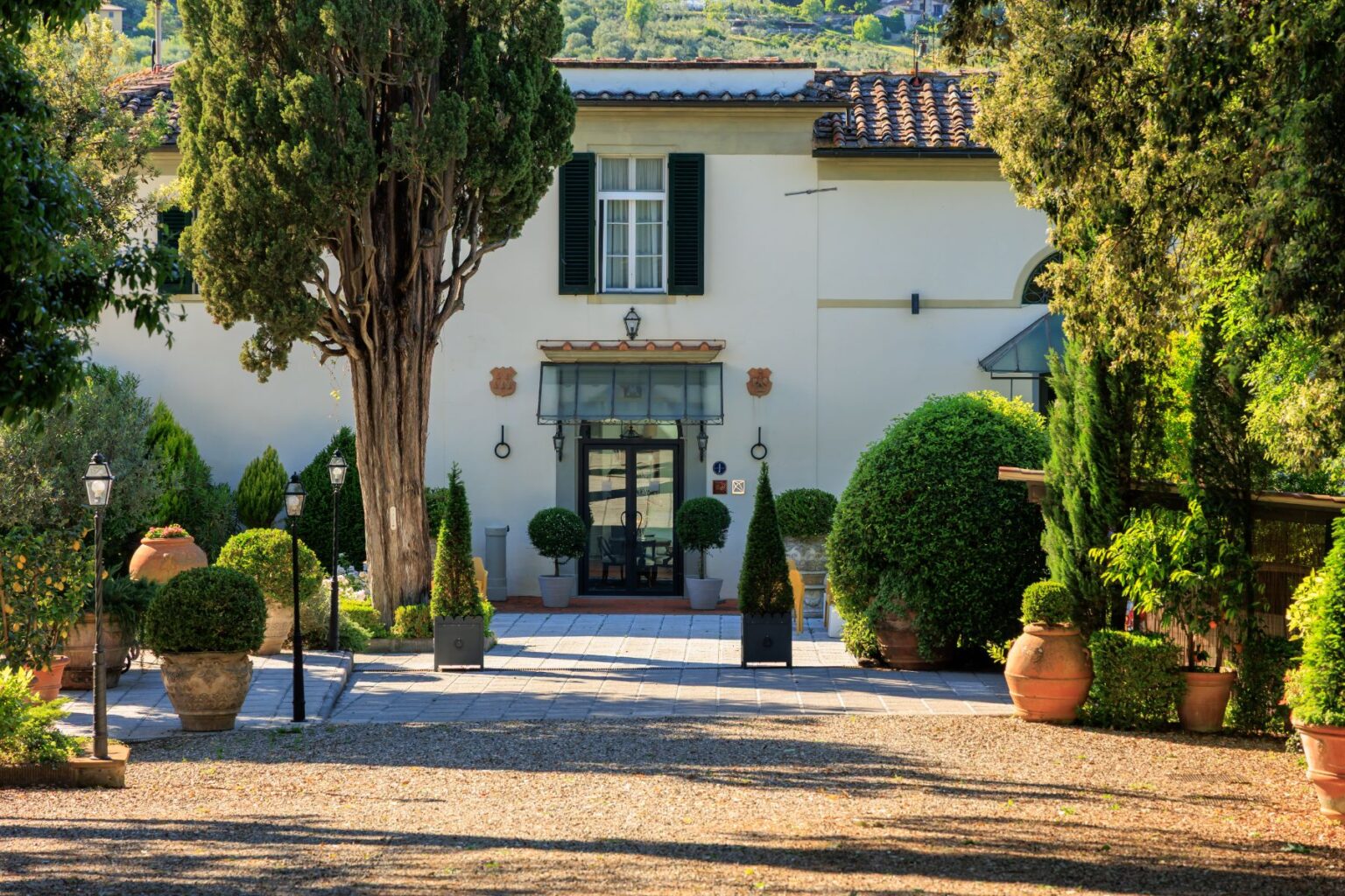 Elegant villa entrance surrounded by lush greenery, including topiary and ornamental trees, with a stone pathway leading to the main door, framed by decorative potted plants and vintage street lamps. Ideal for showcasing luxury accommodations or garden design. | Only on SopranoVillas