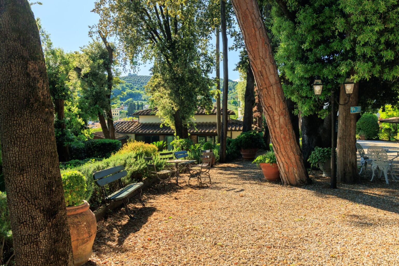 Lush garden pathway with benches and tables, surrounded by trees and greenery, featuring a scenic view of distant hills and rustic buildings, ideal for relaxation and outdoor gatherings. | Only on SopranoVillas