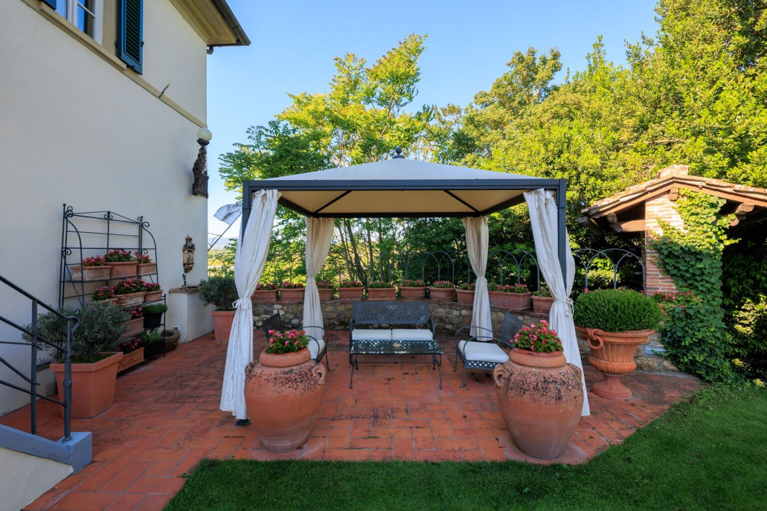 Charming outdoor patio featuring a gazebo with draped curtains, surrounded by potted flowers and terracotta planters, set against a backdrop of lush greenery and a sunny blue sky. Ideal for relaxation and entertaining. | Only on SopranoVillas