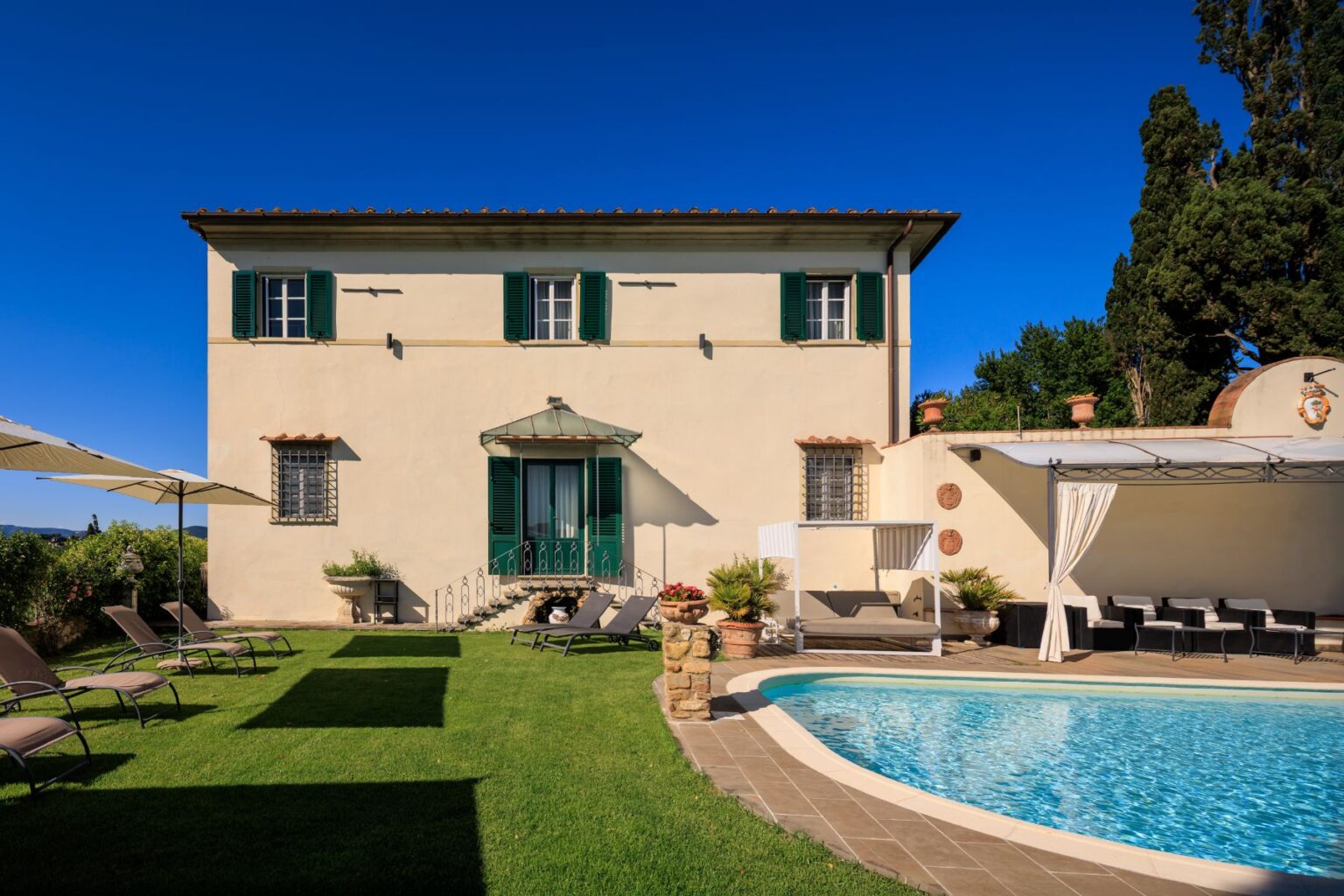 Elegant Tuscan villa with green shutters, surrounded by lush gardens and a swimming pool, featuring sun loungers and a shaded seating area, under a clear blue sky. Perfect for luxury vacations and relaxation. | Only on SopranoVillas