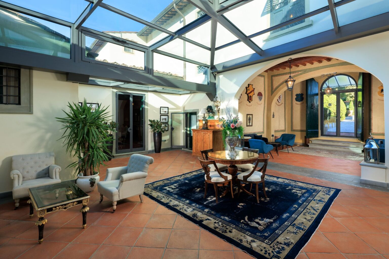 Bright and airy interior of a stylish living space featuring a glass ceiling, elegant furniture, a round dining table, and decorative plants. The room showcases a blend of modern and classic design elements, with terracotta flooring and an inviting atmosphere. | Only on SopranoVillas