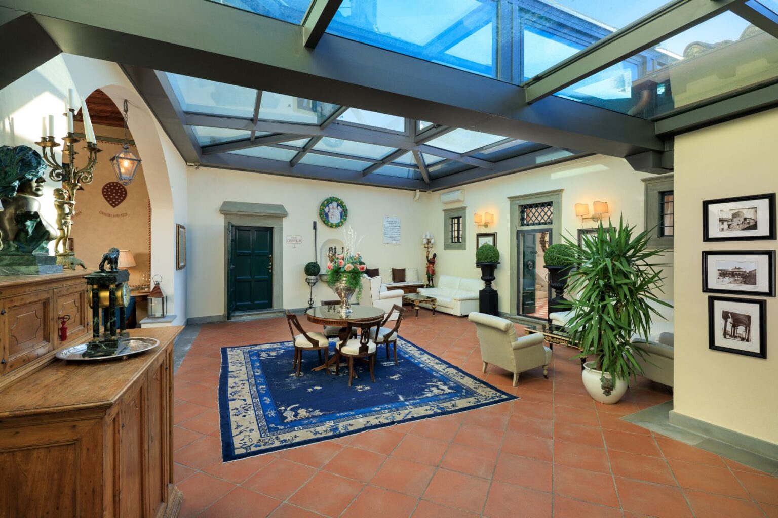 Spacious and elegantly designed living area featuring a glass ceiling, wooden furniture, a round dining table, and decorative plants. The room is adorned with artwork and a large blue area rug, creating a warm and inviting atmosphere. | Only on SopranoVillas