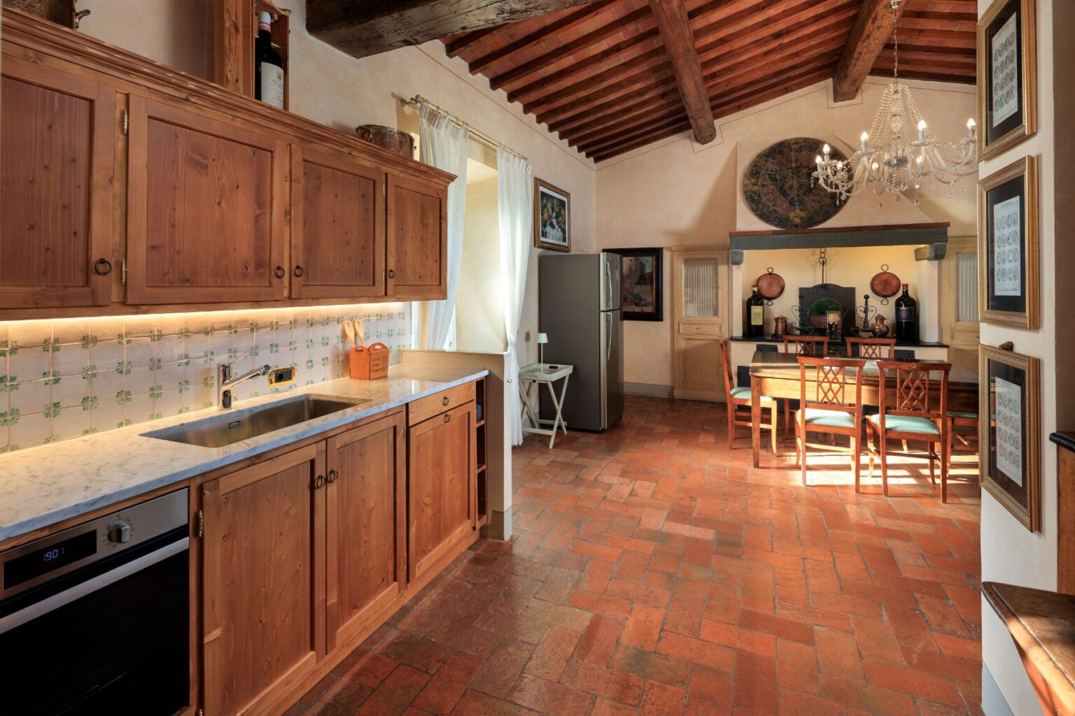 Cozy rustic kitchen featuring wooden cabinets, marble countertops, and terracotta flooring. The space includes modern appliances, a dining area with a wooden table and chairs, and decorative elements like artwork and a chandelier. Natural light illuminates the room, enhancing its warm ambiance. | Only on SopranoVillas