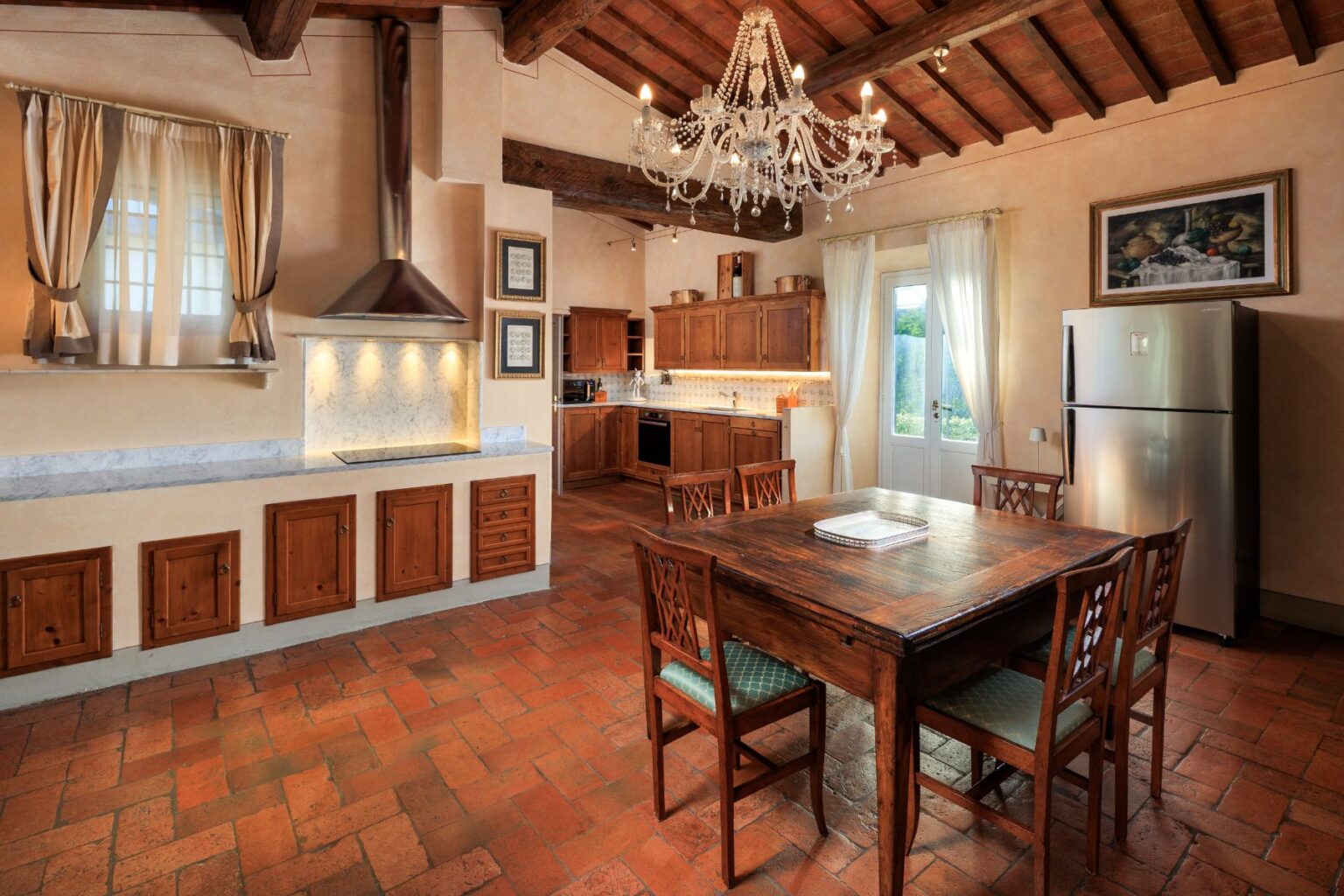 Spacious rustic kitchen with wooden cabinets, marble countertops, and a chandelier, featuring a dining table and modern appliances, bathed in natural light from large windows. | Only on SopranoVillas
