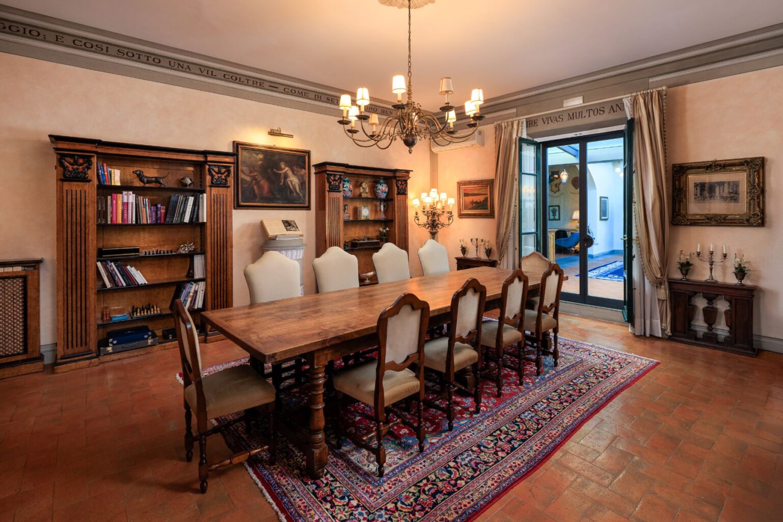 Elegant dining room featuring a large wooden table surrounded by upholstered chairs, flanked by ornate bookshelves filled with books and decorative items. A stylish chandelier hangs overhead, while large doors lead to an outdoor area, enhancing the spacious ambiance. The room is adorned with classic artwork and a richly patterned rug, creating a warm and inviting atmosphere. | Only on SopranoVillas