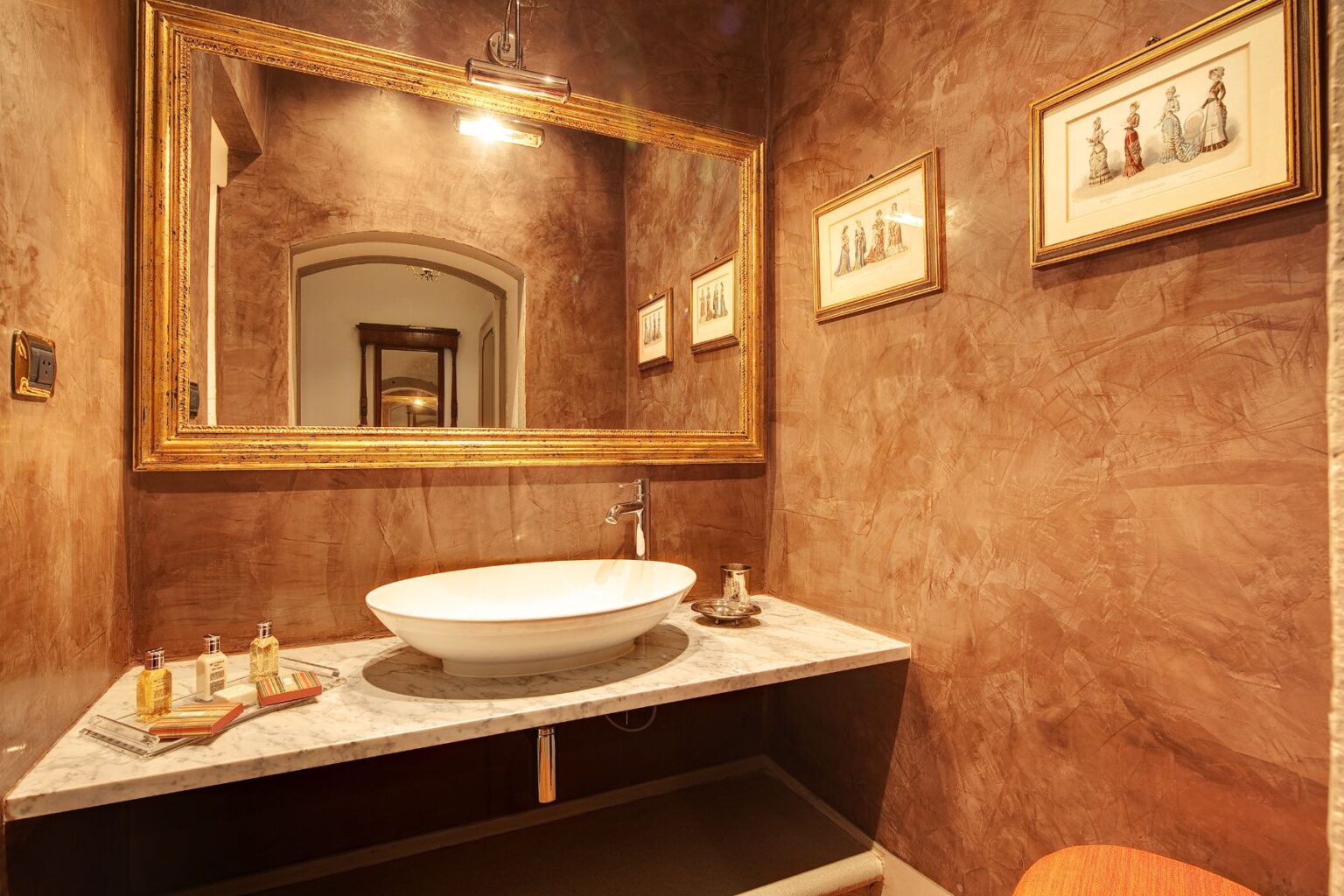 Elegant bathroom featuring a marble countertop with a modern basin, framed mirror, and vintage artwork on textured brown walls, creating a stylish and sophisticated ambiance. | Only on SopranoVillas