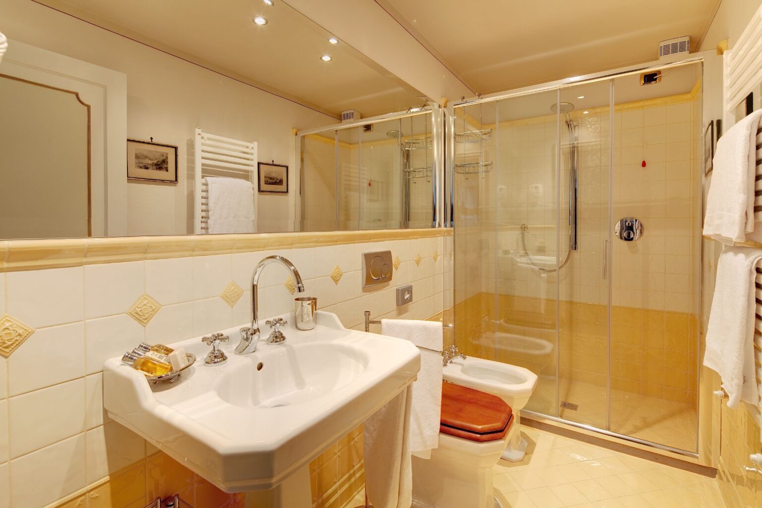 Modern bathroom interior featuring a glass shower enclosure, white sink with chrome fixtures, and elegant yellow and white tiled walls. The space includes a heated towel rack and neatly arranged towels, creating a clean and inviting atmosphere. | Only on SopranoVillas