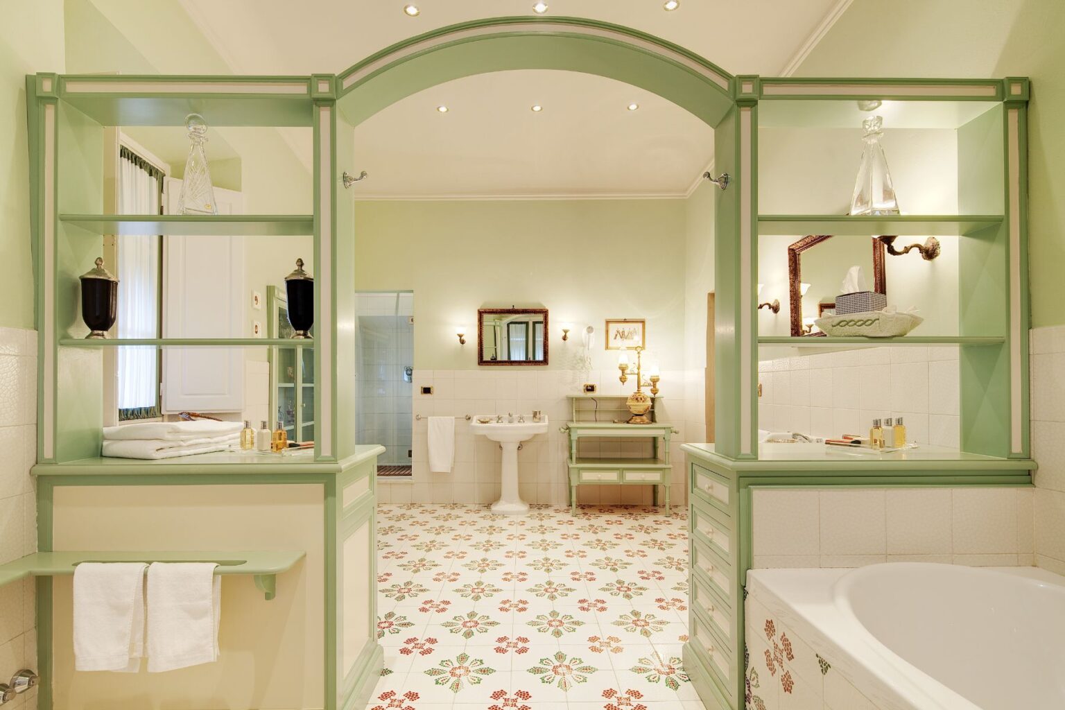 Elegant bathroom featuring light green cabinetry, decorative shelving with glass accents, and a floral tiled floor. The space includes a classic sink, a bathtub, and stylish lighting, creating a serene and inviting atmosphere. Ideal for home improvement or interior design inspiration. | Only on SopranoVillas