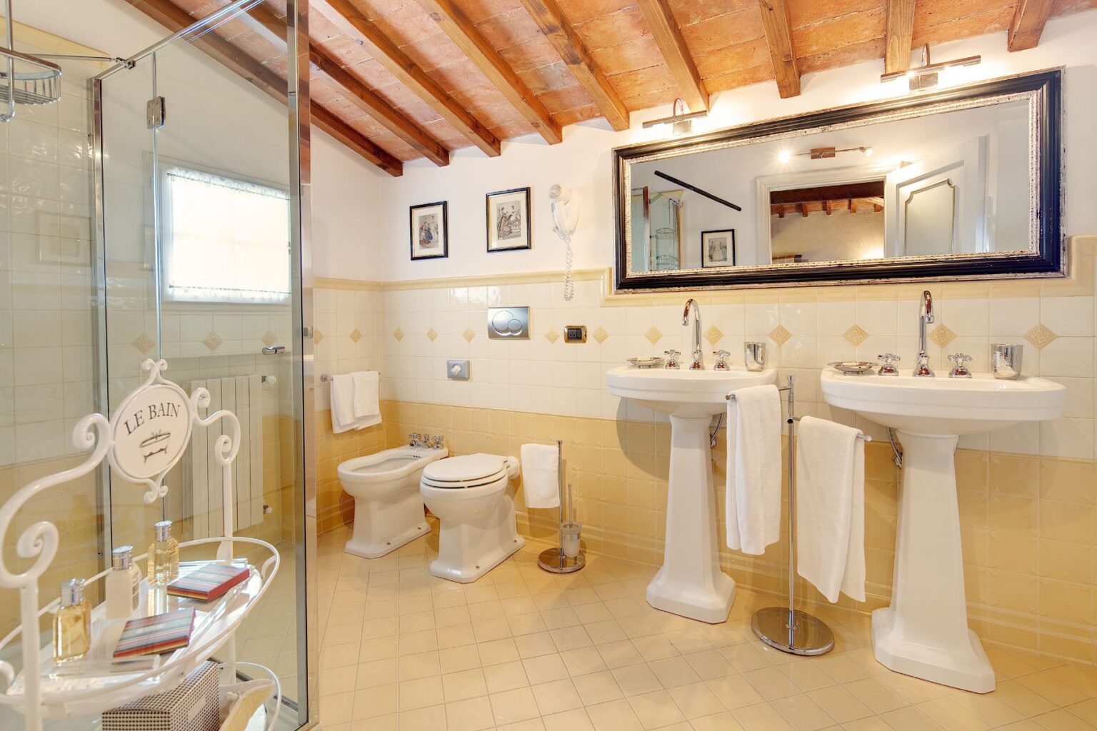 Stylish bathroom featuring a glass shower, double sinks, and elegant decor with wooden beams and yellow tiles. The space is well-lit and includes toiletries on a decorative stand, creating a luxurious and inviting atmosphere. | Only on SopranoVillas