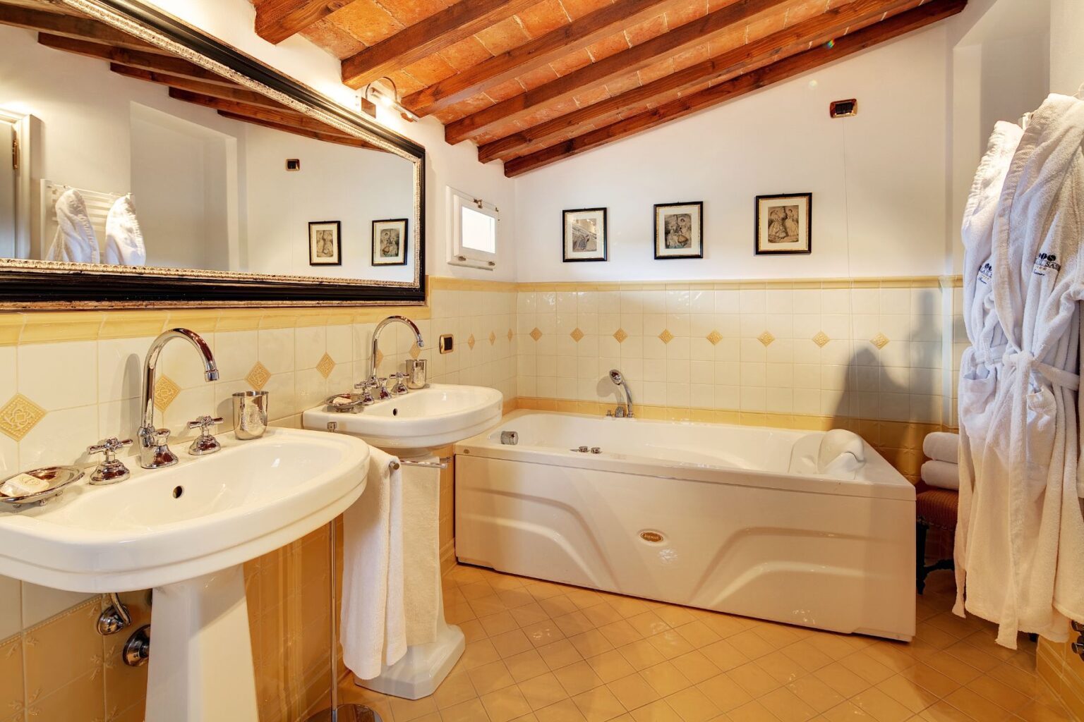 Bright and elegant bathroom featuring double sinks, a large bathtub, and a stylish mirror. The room showcases wooden beams, tiled walls with decorative accents, and framed artwork, creating a cozy and inviting atmosphere. Fluffy white robes and neatly arranged towels add a touch of luxury. | Only on SopranoVillas