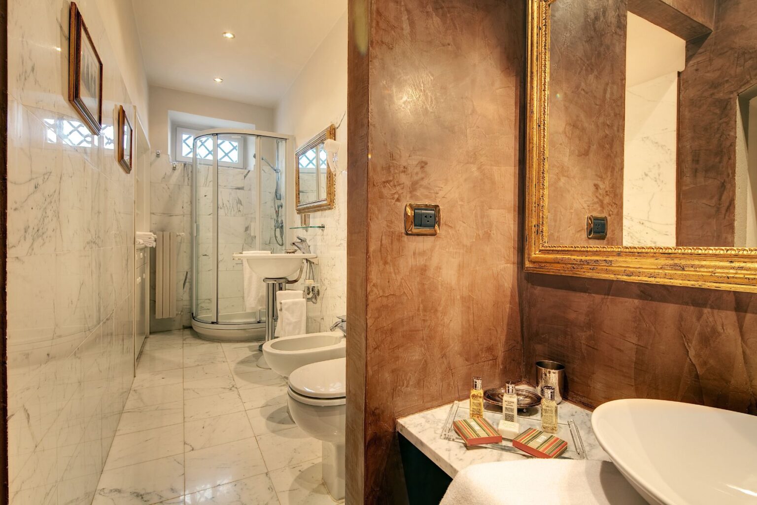 Modern bathroom featuring marble walls and flooring, a glass shower enclosure, a stylish sink with decorative toiletries, and elegant framed mirrors, creating a luxurious and inviting atmosphere. | Only on SopranoVillas