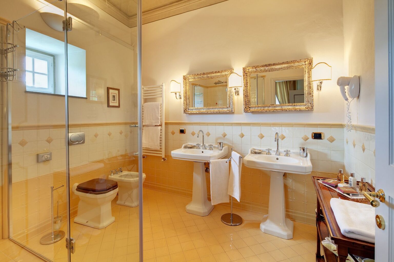 Luxurious bathroom featuring dual pedestal sinks, a glass shower enclosure, and elegant decor. The space is accented with yellow tiles, framed mirrors, and a stylish wooden vanity with toiletries, creating a bright and inviting atmosphere. | Only on SopranoVillas