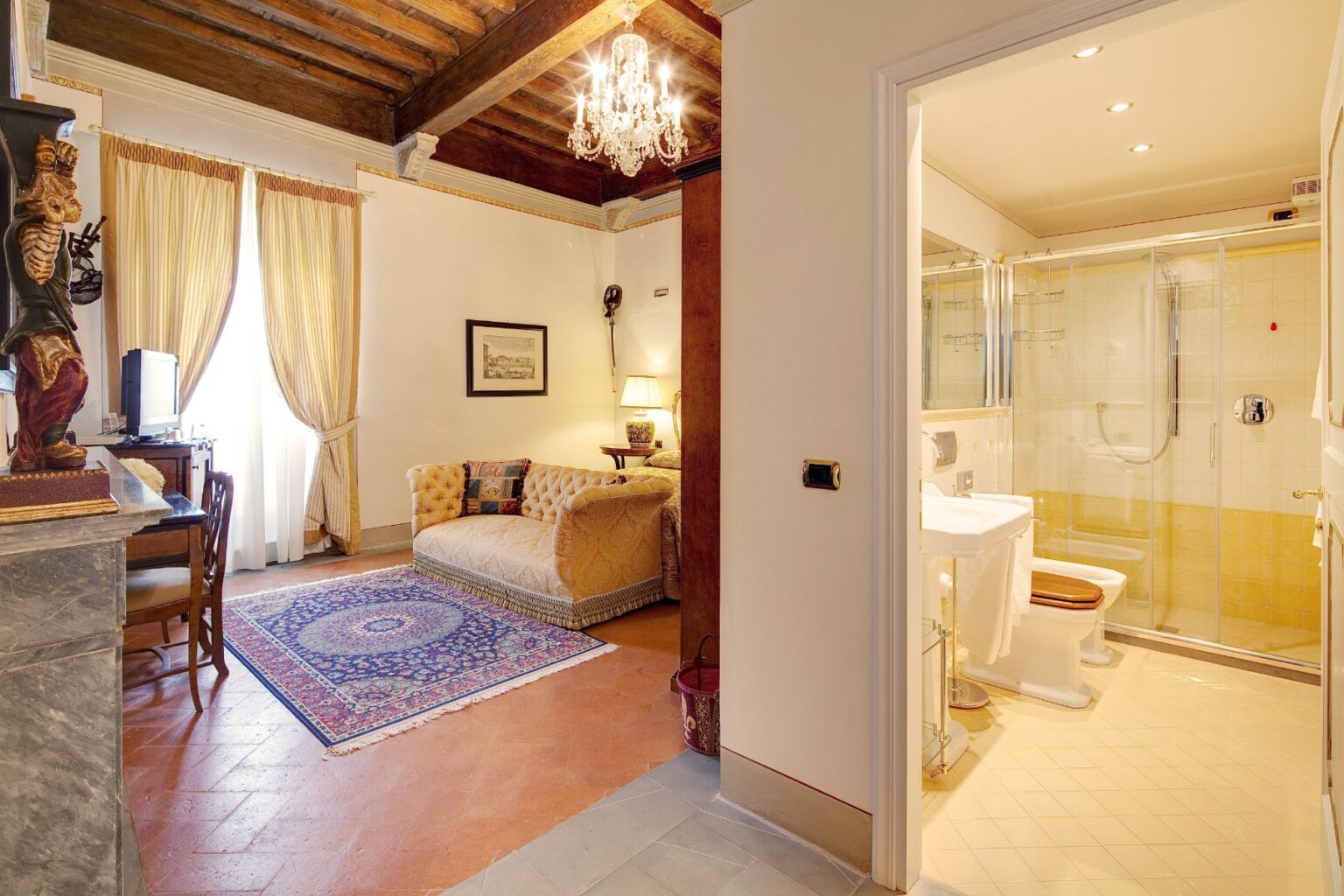 Cozy hotel room featuring a plush sofa, elegant decor, and a small workspace, with an adjoining bathroom showcasing modern fixtures and a glass shower. The space is illuminated by natural light from large windows and a chandelier, highlighting the charming interior design. | Only on SopranoVillas