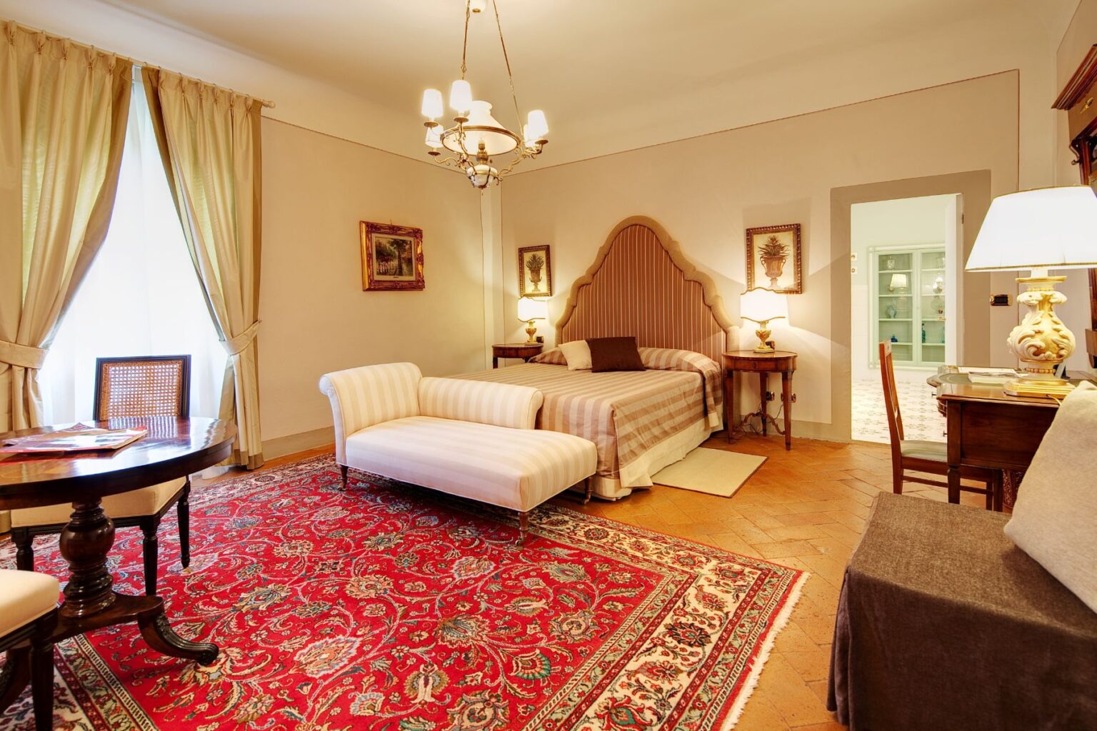 Elegant hotel room featuring a king-sized bed with a striped headboard, a plush chaise lounge, and a wooden dining table. The space is adorned with warm lighting, decorative curtains, and a vibrant red area rug, creating a cozy and inviting atmosphere. | Only on SopranoVillas