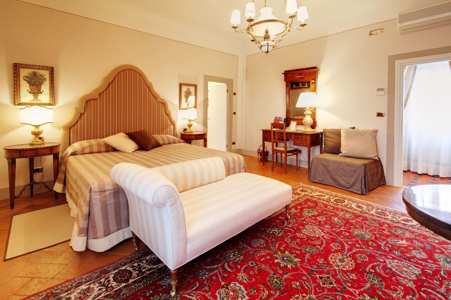 Elegant hotel room featuring a large striped bed with a decorative headboard, a plush white chaise lounge, and wooden furniture. The room is adorned with warm lighting, a vintage writing desk, and a vibrant red patterned rug, creating a cozy and inviting atmosphere. | Only on SopranoVillas