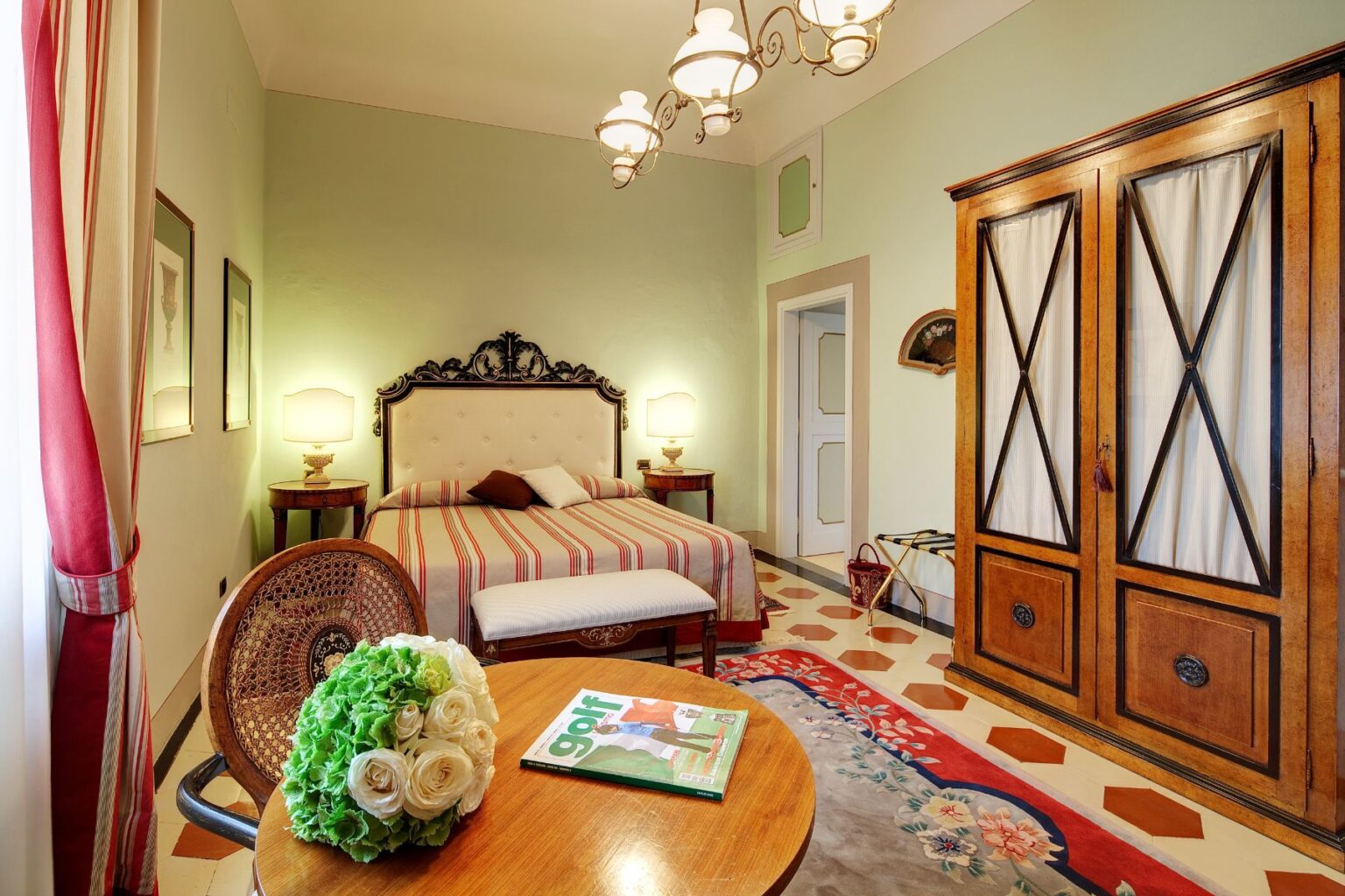 Cozy bedroom featuring a vintage style with a striped bedspread, elegant headboard, and decorative lamp. A wooden wardrobe and a small round table with a floral arrangement and a golf magazine complete the inviting atmosphere. The room showcases soft green walls and patterned flooring, creating a warm and welcoming environment. | Only on SopranoVillas