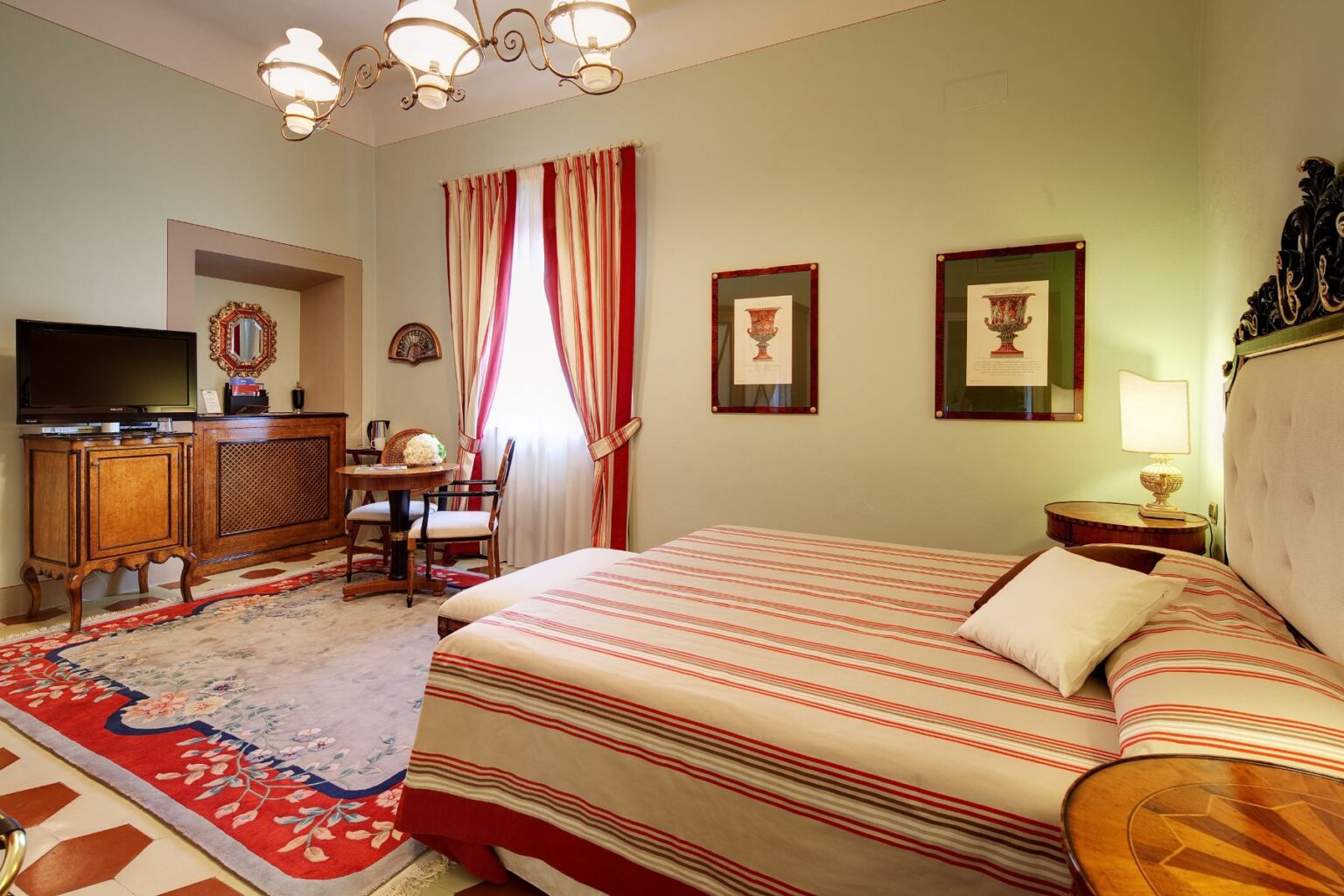 Cozy hotel room featuring a double bed with striped bedding, elegant wooden furniture, a vintage TV, and a small dining area. The room is adorned with framed artwork and has large windows with striped curtains, creating a warm and inviting atmosphere. | Only on SopranoVillas