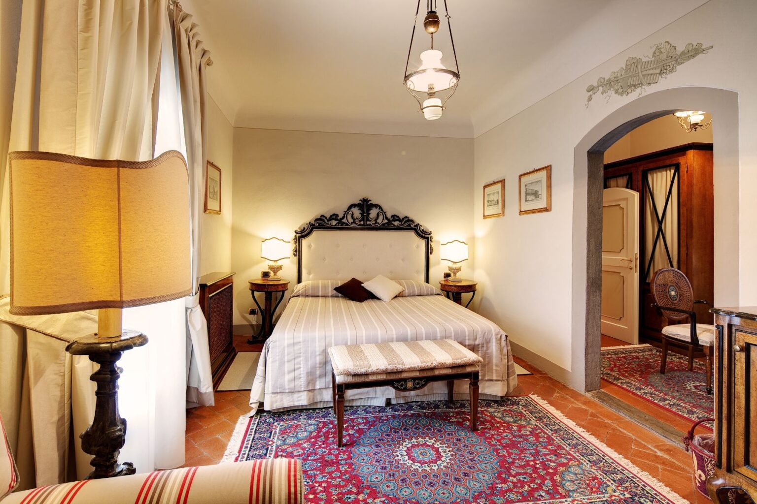 Elegant bedroom interior featuring a plush bed with decorative pillows, vintage lamps, and a cozy seating area. The room showcases a rich patterned rug, warm lighting, and classic decor elements, creating a welcoming atmosphere. Ideal for relaxation and comfort. | Only on SopranoVillas
