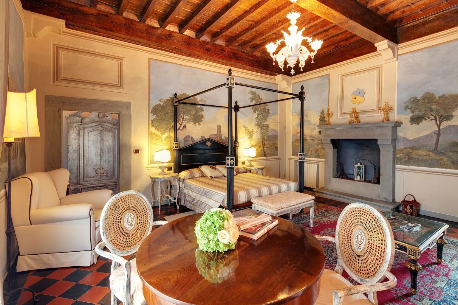Elegant bedroom featuring a four-poster bed, vintage furnishings, and beautiful mural paintings. The room includes a cozy seating area, a wooden table with floral decor, and a decorative fireplace, all set beneath a charming chandelier. | Only on SopranoVillas