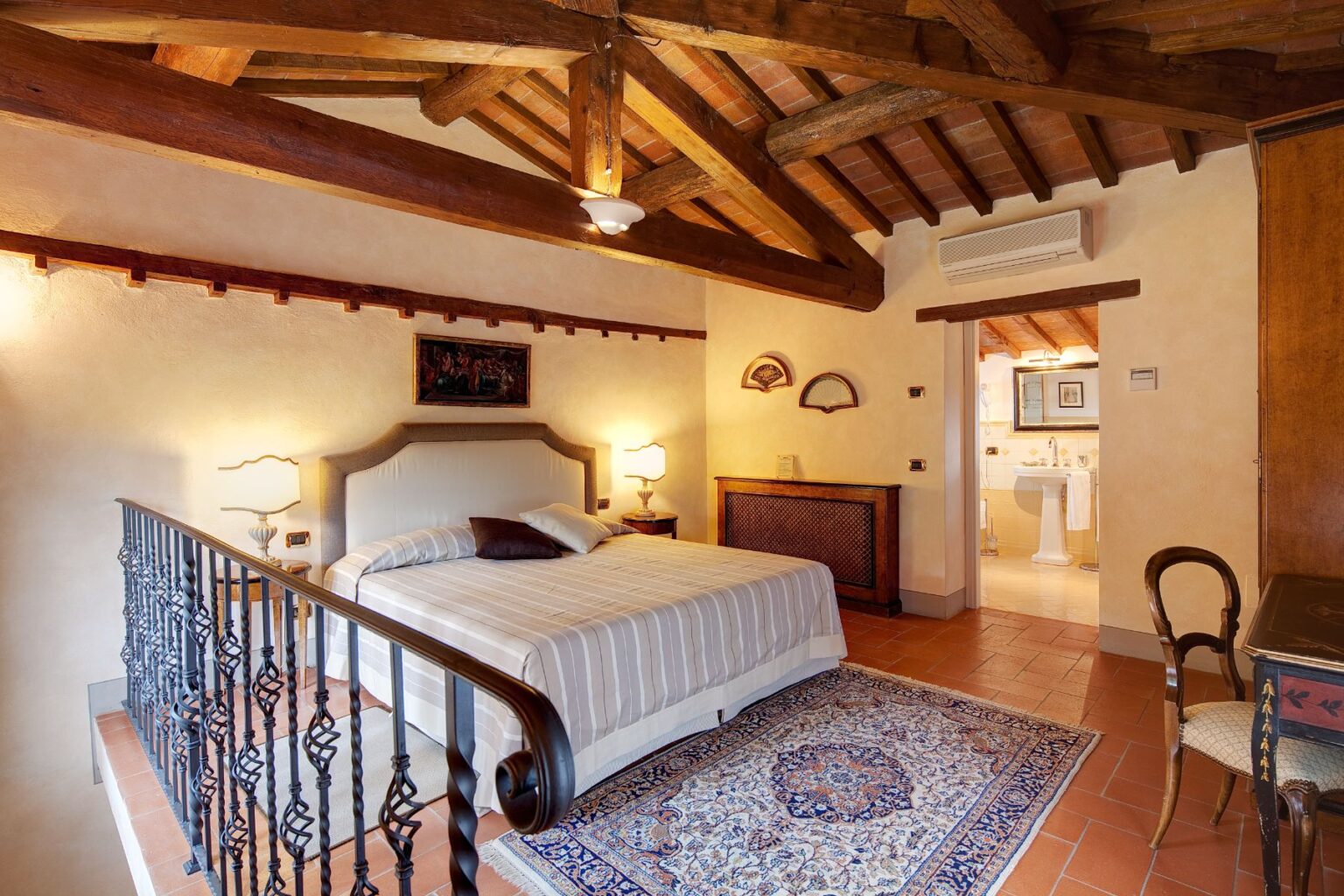 Cozy bedroom featuring a king-sized bed with a striped duvet, wooden beams on the ceiling, and a decorative rug. The room includes warm lighting, a vintage wooden dresser, and an attached modern bathroom visible in the background. Ideal for a relaxing getaway. | Only on SopranoVillas