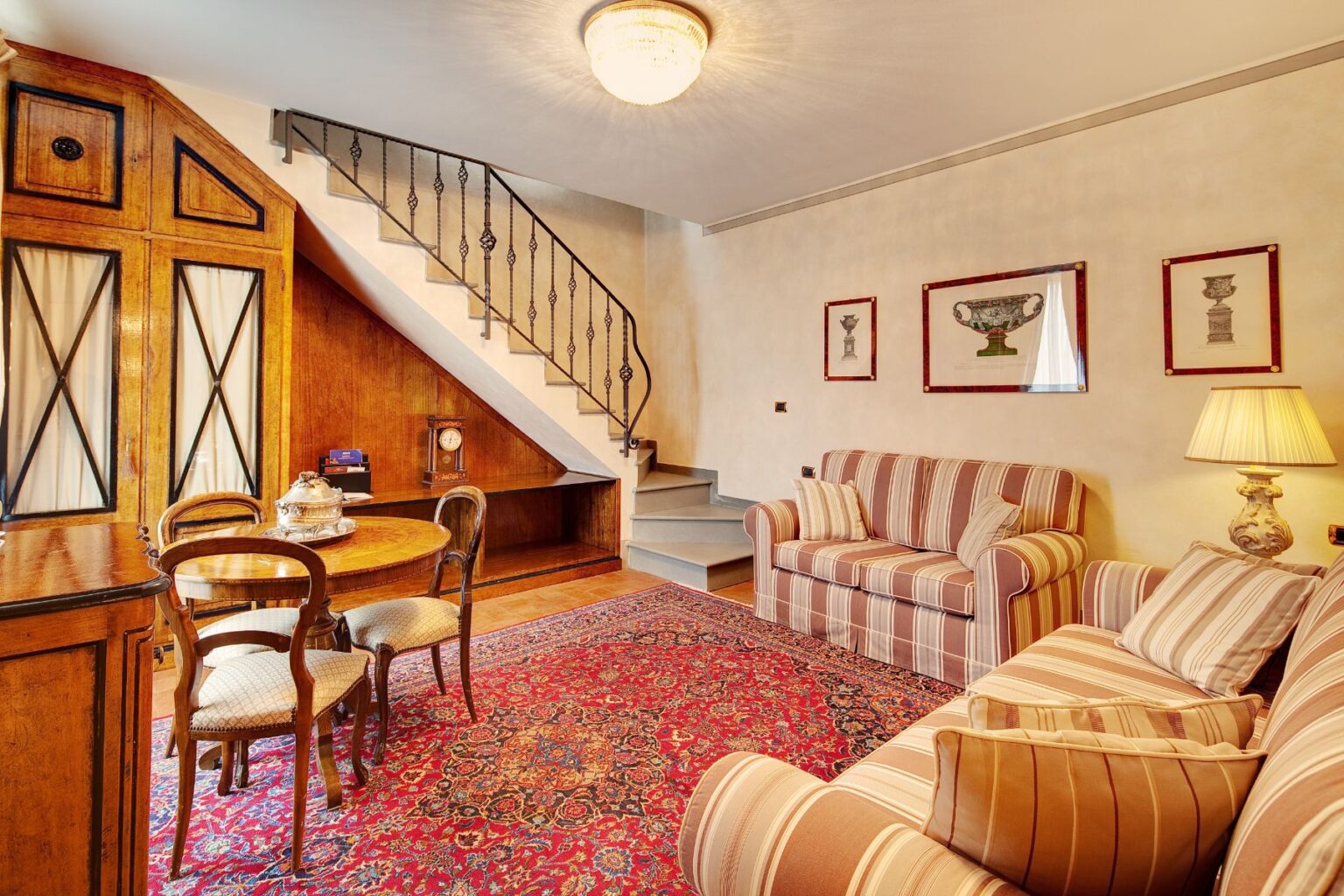 Cozy living room featuring a striped sofa, elegant wooden dining table with chairs, and decorative wall art. The space includes a stylish chandelier and staircase, complemented by a patterned rug, creating a warm and inviting atmosphere. | Only on SopranoVillas