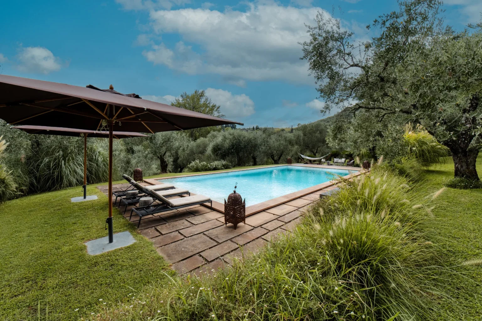 Scenic outdoor swimming pool surrounded by lush greenery and olive trees, featuring lounge chairs under a large umbrella and a decorative lantern on the patio, ideal for relaxation and leisure. | Only on SopranoVillas