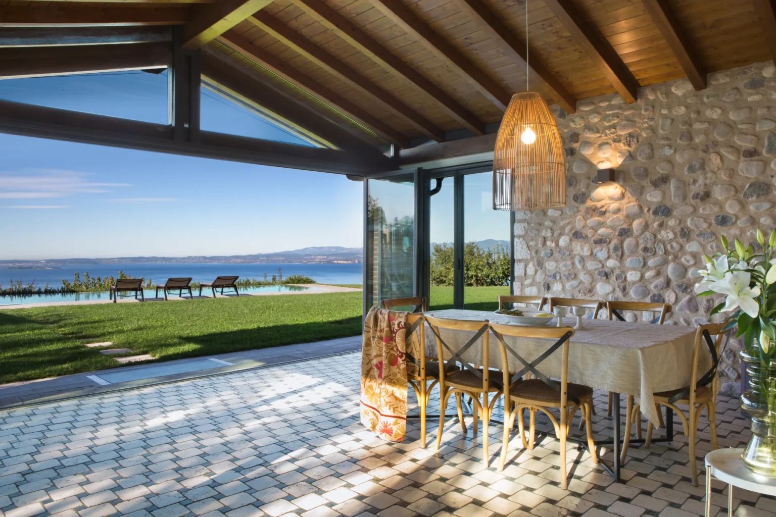 Modern dining area with a stone wall and wooden ceiling, featuring a table set for meals, surrounded by chairs. Large glass doors open to a scenic view of a lush garden and swimming pool overlooking the water and mountains in the background. Natural light floods the space, highlighted by a stylish pendant light. | Only on SopranoVillas
