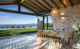 Modern dining area with a stone wall and wooden ceiling, featuring a table set for meals, surrounded by chairs. Large glass doors open to a scenic view of a lush garden and swimming pool overlooking the water and mountains in the background. Natural light floods the space, highlighted by a stylish pendant light.