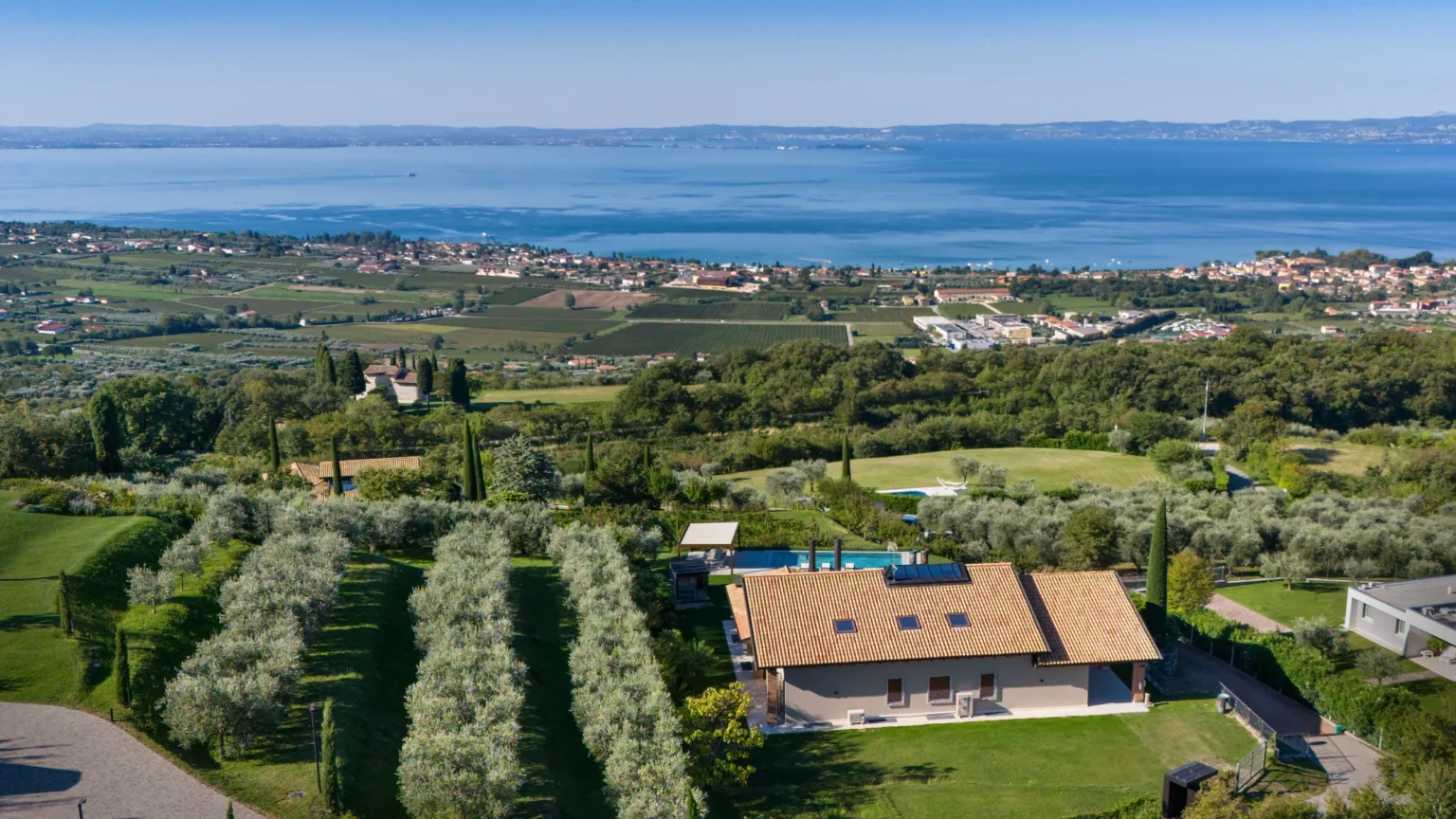 Aerial view of a modern villa surrounded by lush greenery and olive trees, overlooking a serene lake and picturesque landscape with vineyards and residential areas in the background. Ideal for luxury retreats or vacation rentals. | Only on SopranoVillas
