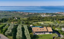 Aerial view of a modern villa surrounded by lush greenery and olive trees, overlooking a serene lake and picturesque landscape with vineyards and residential areas in the background. Ideal for luxury retreats or vacation rentals.