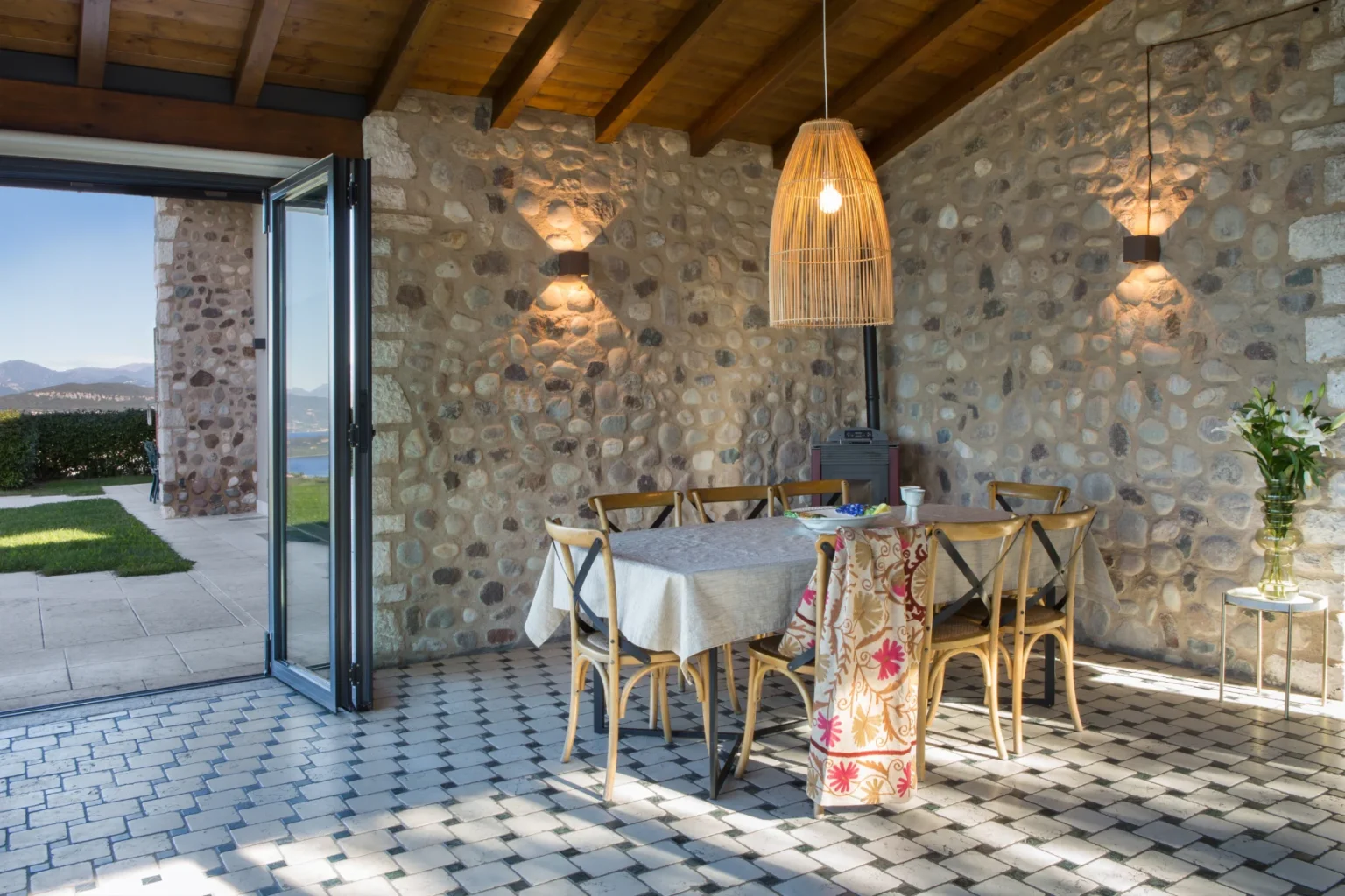 Cozy dining area with a stone wall, wooden ceiling, and modern decor, featuring a large table surrounded by chairs, a stylish pendant light, and a view of the outdoor garden and mountains. Ideal for inviting gatherings and meals. | Only on SopranoVillas