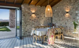 Cozy dining area with a stone wall, wooden ceiling, and modern decor, featuring a large table surrounded by chairs, a stylish pendant light, and a view of the outdoor garden and mountains. Ideal for inviting gatherings and meals.