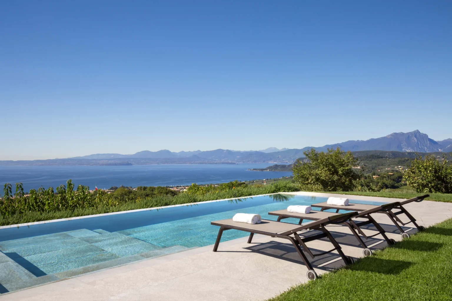 Stunning view of a modern infinity pool with lounge chairs overlooking a scenic lake and mountainous landscape under a clear blue sky. Ideal for luxury vacation rentals or serene getaways. | Only on SopranoVillas