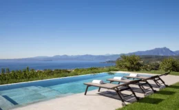 Stunning view of a modern infinity pool with lounge chairs overlooking a scenic lake and mountainous landscape under a clear blue sky. Ideal for luxury vacation rentals or serene getaways.