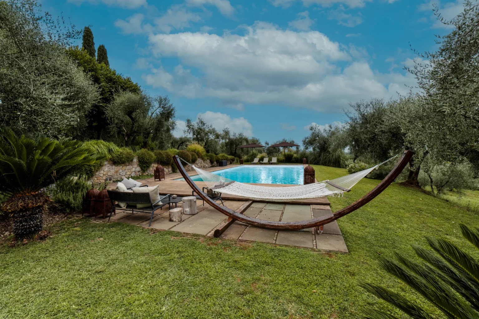 Scenic outdoor space featuring a hammock by a sparkling pool surrounded by lush greenery, including olive trees and tropical plants, ideal for relaxation and leisure. | Only on SopranoVillas