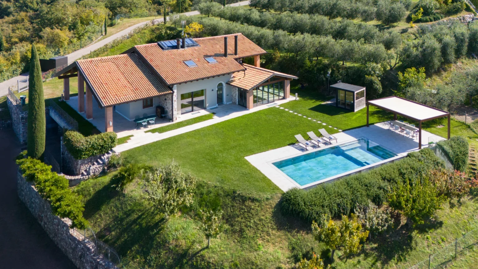 Aerial view of a modern villa with a tiled roof, surrounded by lush greenery and olive trees. The property features a spacious lawn, a swimming pool with lounge chairs, and a shaded outdoor dining area, showcasing a tranquil and luxurious countryside retreat. | Only on SopranoVillas