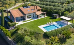 Aerial view of a modern villa with a tiled roof, surrounded by lush greenery and olive trees. The property features a spacious lawn, a swimming pool with lounge chairs, and a shaded outdoor dining area, showcasing a tranquil and luxurious countryside retreat.